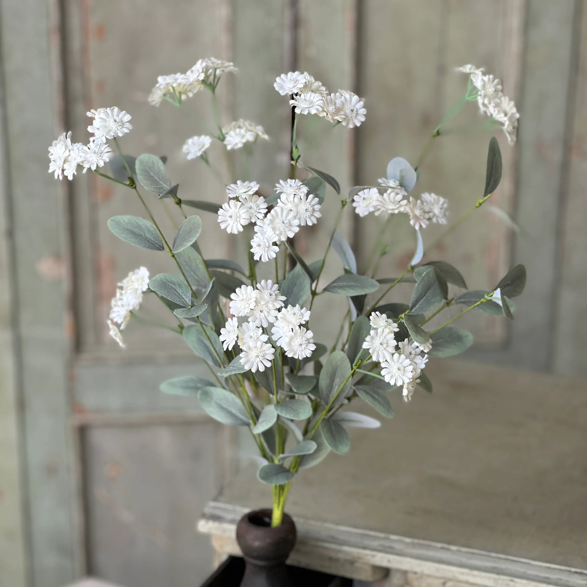 Faux floral arrangement in a black pot on a decorative wooden surface with a rustic background.